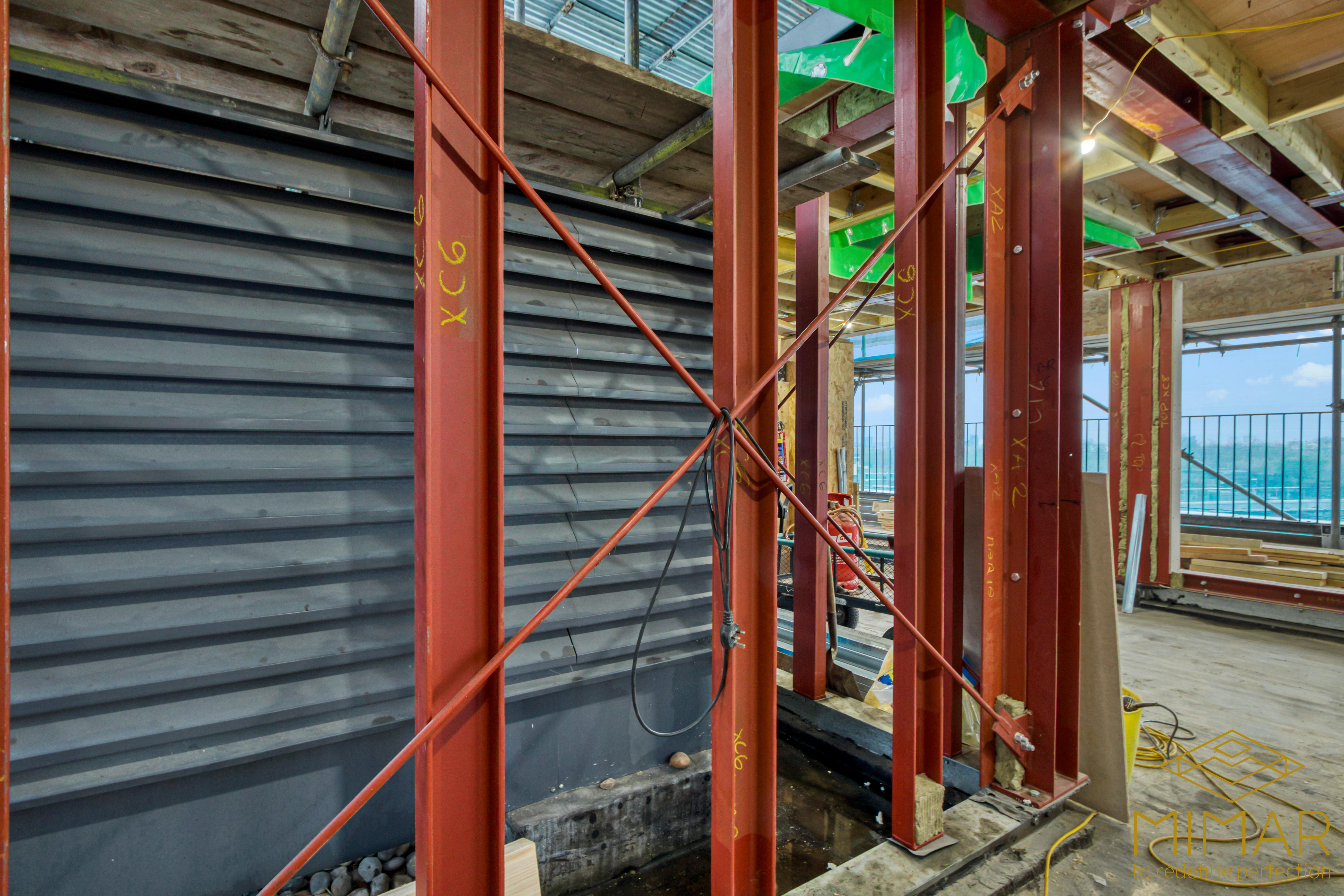 The image shows a partially constructed building with exposed red steel beams connected by cross bracing, indicating structural support within a new build construction site.